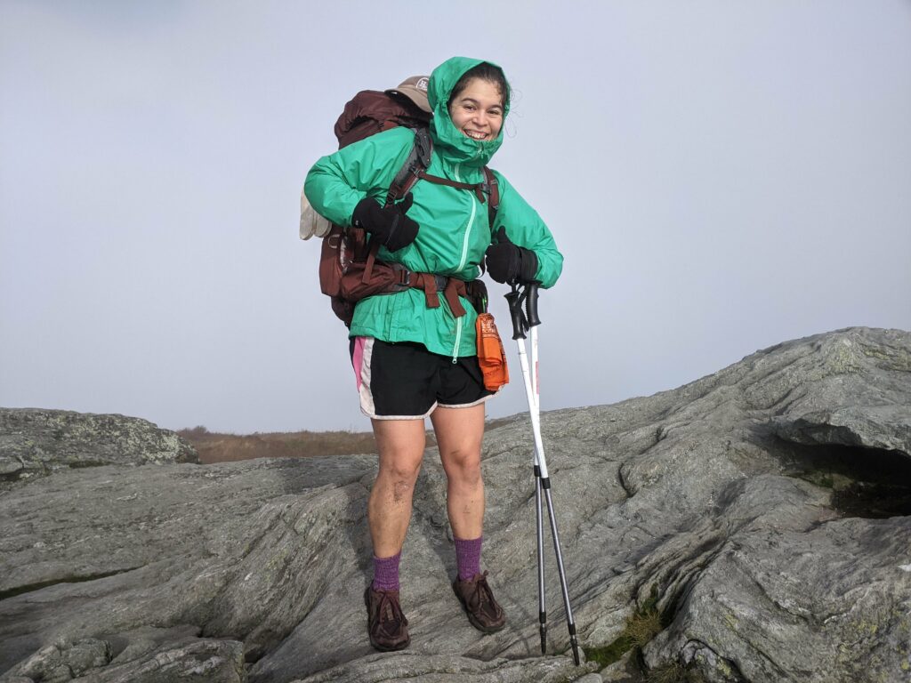 A woman in a green rain coat, black shorts, hiking socks and shoes, black mittens, trekking poles, and a maroon backpack smiling and giving two thumbs up. She is standing on open rock face and the background is all foggy clouds